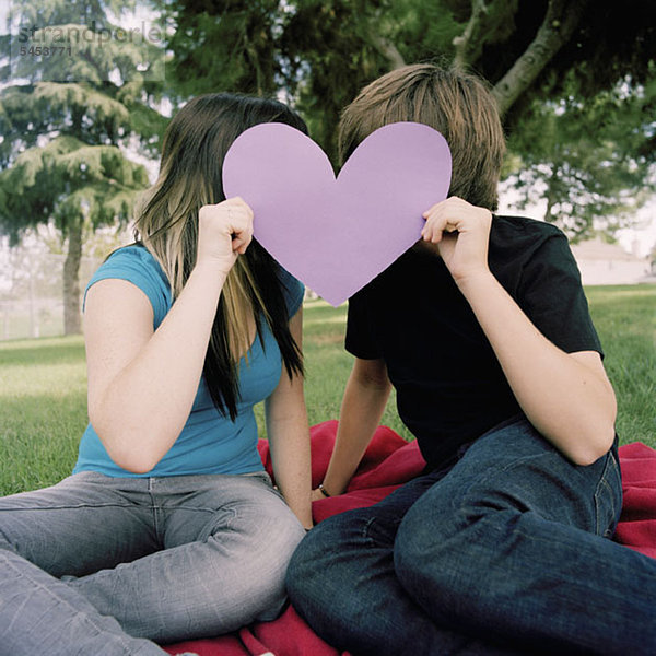 Ein junges Paar  das sich hinter einem ausgeschnittenen Herzen versteckt.