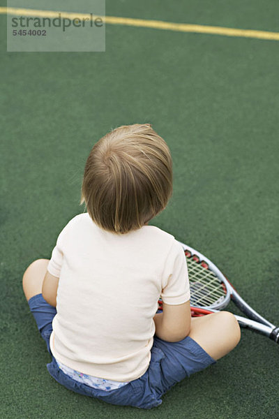 Ein kleiner Junge sitzt auf einem Tennisplatz mit einem Tennisschläger  Rückansicht