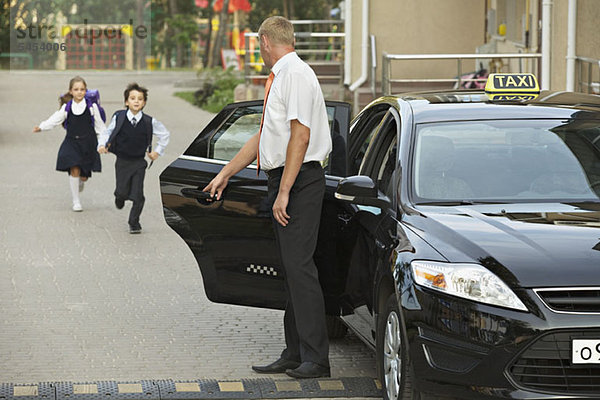 Zwei Kinder in Schuluniformen laufen auf das schwarze Taxi zu.