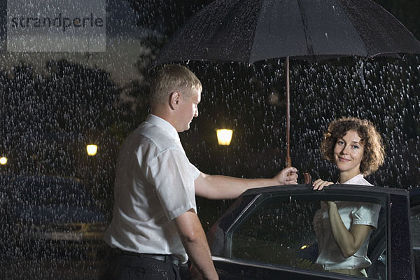 Ein Mann öffnet eine Autotür und hält einen Regenschirm über eine Frau.