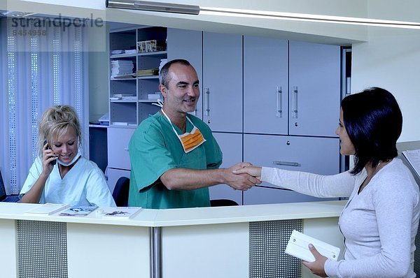 Patientin begrüßt Arzt mit Handschlag an der Rezeption von einer Arztpraxis - Im Hintergrund eine Helferin am Telefon