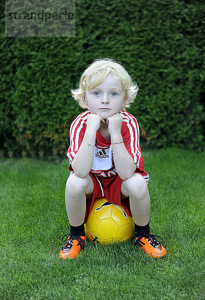 Fünfjähriger Junge in FC Bayern München Dress sitzt auf gelbem Fußball