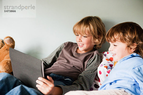 Zwei kleine Jungen auf einem Bett  die ein digitales Tablett benutzen.