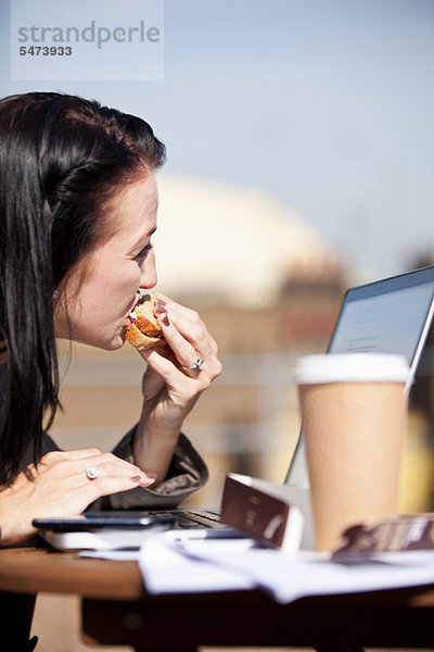 Junge Frau isst Sandwich mit Laptop