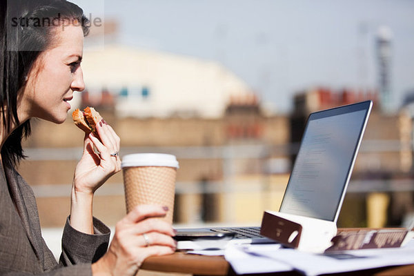 Junge Frau  die draußen einen Laptop benutzt  während sie ein Sandwich isst.