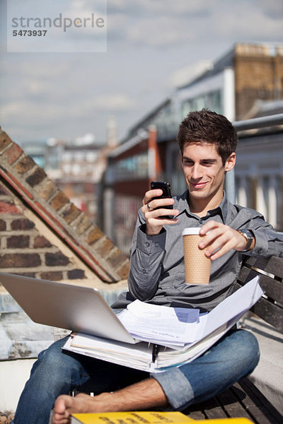Junger Mann im Dachgarten mit Laptop und Handy