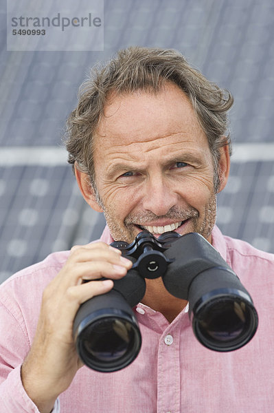 Erwachsener Mann hält Fernglas in Solaranlage  lächelnd  Portrait