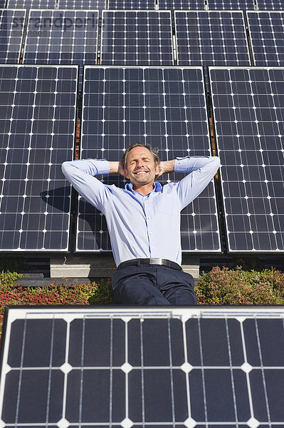 Reifer Mann  der sich auf einem Panel in einer Solaranlage ausruht  lächelnd