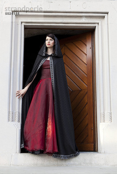Junge Frau mit rotem Kleid und schwarzem Überhang steht an Tür