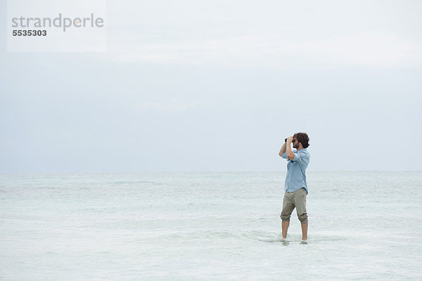 Mann im Meer stehend  durchs Fernglas schauend