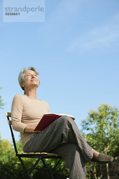 Seniorin im Freien sitzend mit Buch  Sonnenwärme genießend