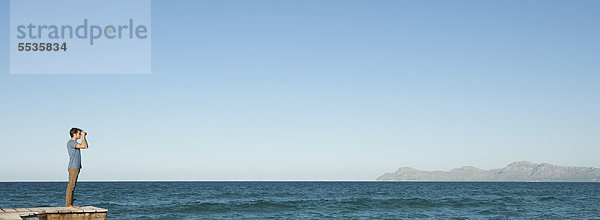 Mann in der Ferne steht auf dem Pier und schaut durch ein Fernglas auf das Meer.
