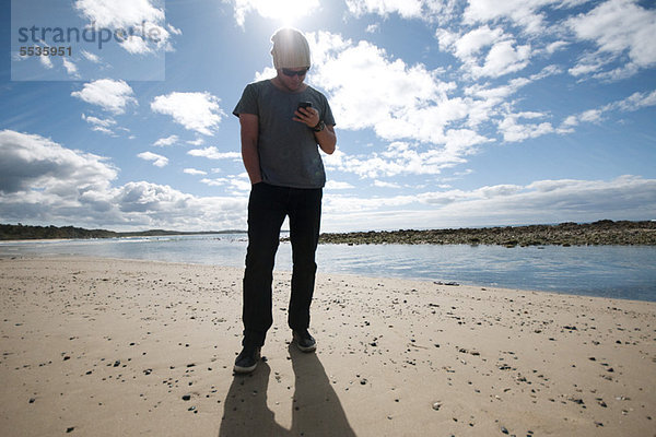 Mann steht am Strand und schaut auf sein Handy.