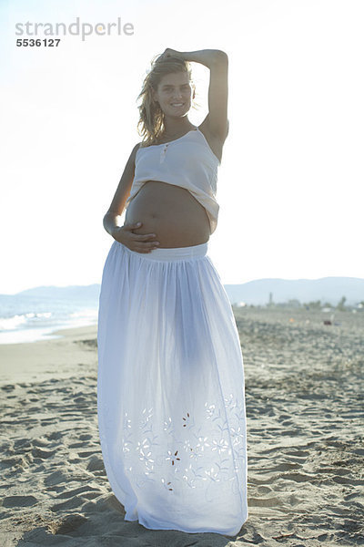 Schwangere Frau am Strand stehend mit Hand am Bauch