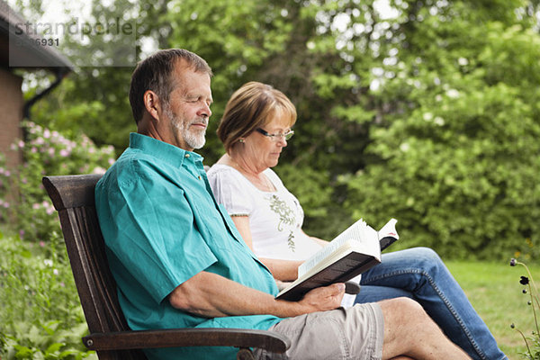 Seniorenpaar liest Buch im Garten  während es im Sessel sitzt.