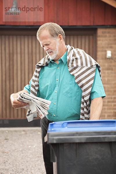 Älterer Mann  der Zeitung liest  steht in der Nähe des Mülleimers.