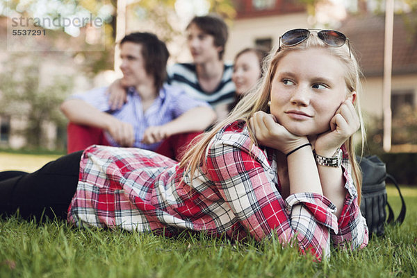 Teenagermädchen auf Gras liegend mit Freunden im Hintergrund