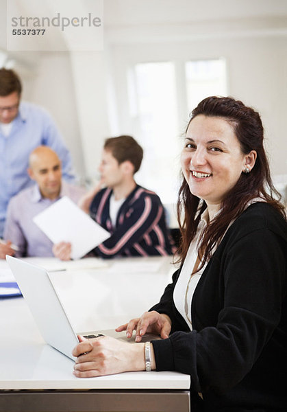 Lächelnde Geschäftsfrau mit Laptop und Kollegen am Konferenztisch im Hintergrund