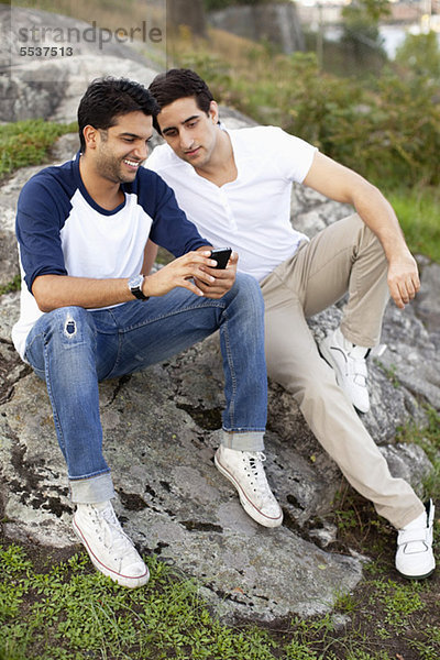 Volle Länge von lächelnden jungen männlichen Freunden  die zusammen das Handy anschauen.