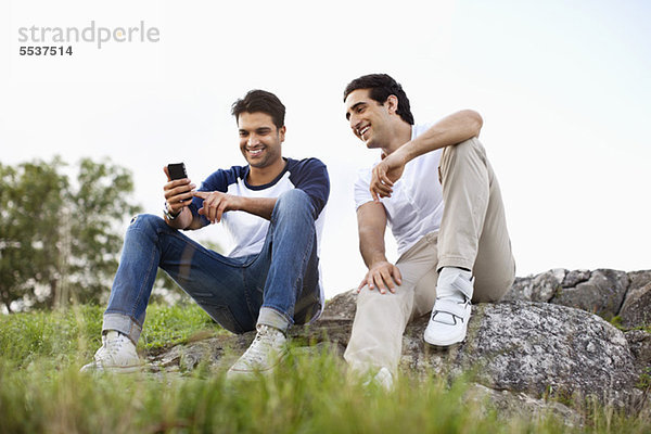 Volle Länge von jungen Freunden  die mit dem Handy auf dem Felsen sitzen.