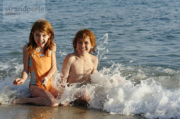 Schwester und Bruder sitzen am Strand im Wasser und freuen sich in den Wellen