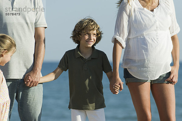 Ein Junge am Strand mit seiner Familie  der die Hände seiner Eltern hält.