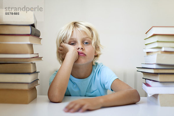 Junge sitzt zwischen zwei Stapeln von Büchern mit gelangweiltem Gesichtsausdruck.