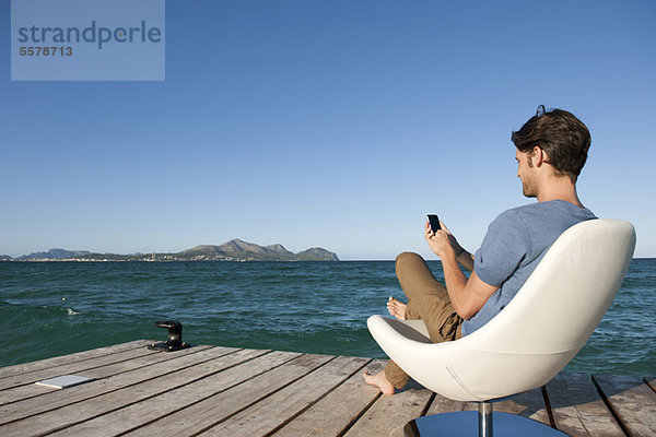 Junger Mann im Sessel am See mit Handy  Seitenansicht