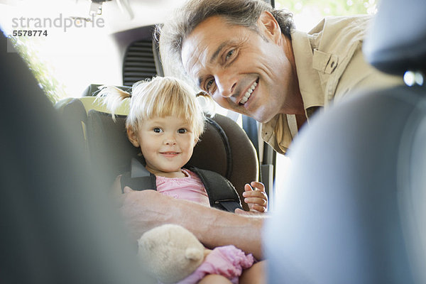 Vater befestigt kleines Mädchen im Autositz  beide lächelnd vor der Kamera