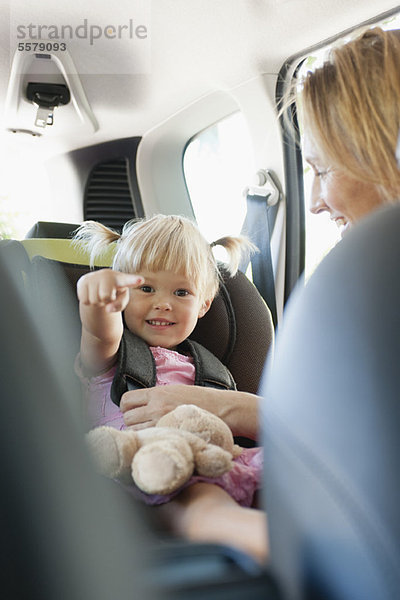 Kleines Mädchen zeigt und lächelt  als ihre Mutter sie im Autositz befestigt.