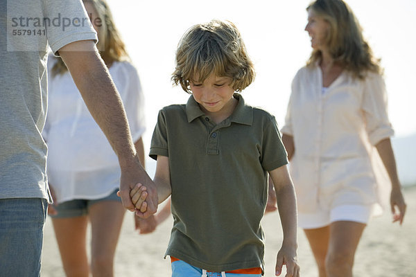 Junge  der mit seiner Familie am Strand spazieren geht und Vaters Hand hält.