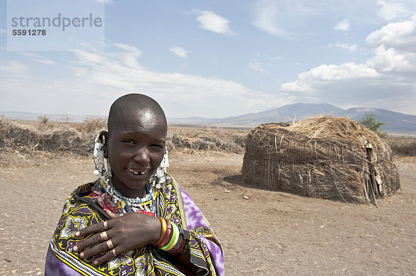 Portrait  Ethnologie  Frau der Massai trägt Schmuck  Maasai  kahl rasierter Kopf  vor Hütte aus Stroh und Ästen  Dorf Kiloki  Savanne  Ngorongoro Conservation Area  Serengeti Nationalpark  Tansania  Ostafrika  Afrika