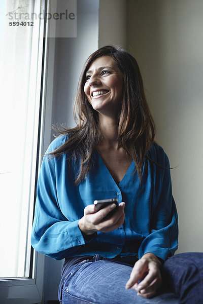 Mittlere erwachsene Frau am Fenster mit Handy  lächelnd