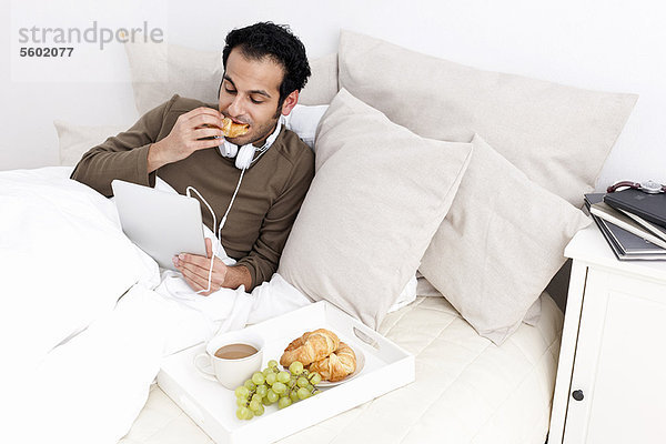 Mann mit Tablet-Computer beim Essen im Bett