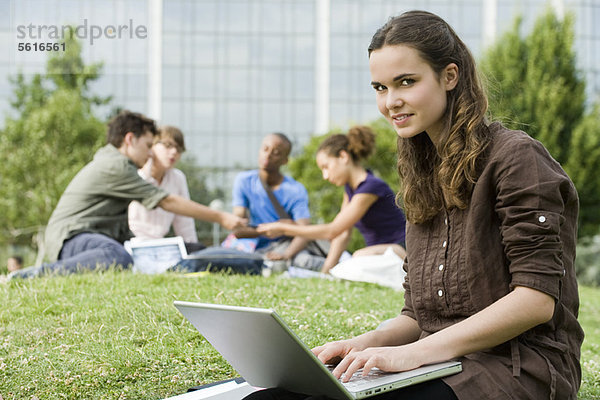 Junge Frau mit Laptop auf Rasen  Menschen im Hintergrund