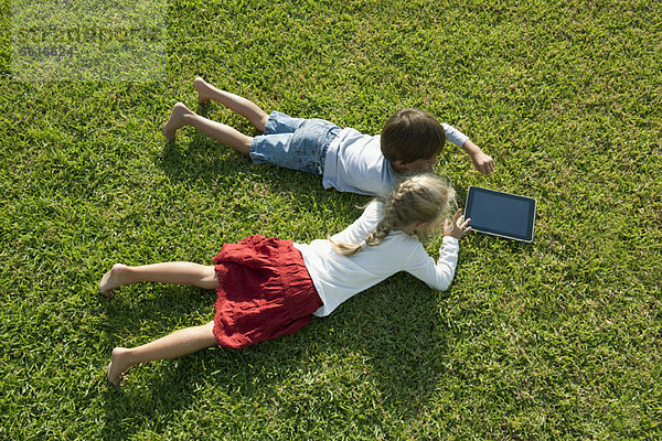 Kinder  die auf Gras liegen  benutzen zusammen ein digitales Tablett.