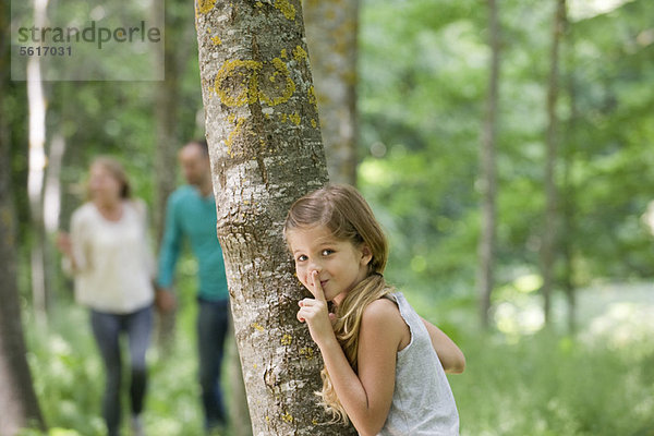 Mädchen versteckt sich hinter Baum mit Finger auf den Lippen