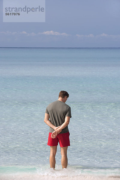 Mann steht knöcheltief im Wasser und betrachtet den Ozean  Rückansicht
