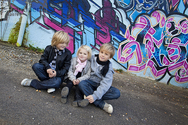 Geschwister  2  5 und 7 Jahre  sitzen vor Graffiti-Wand auf einem Skateboard