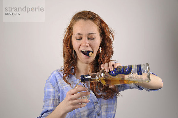 Junge Frau mit roten Haaren öffnet eine Flasche Whiskey mit den Zähnen und gießt sich ein Glas ein