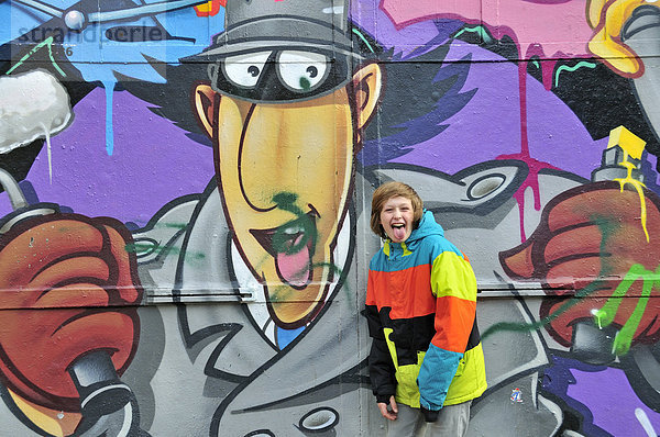 Zwölfjähriger Junge streckt frech die Zunge heraus  vor Graffitiwand  Köln  Nordrhein-Westfalen  Deutschland  Europa  ÖffentlicherGrund