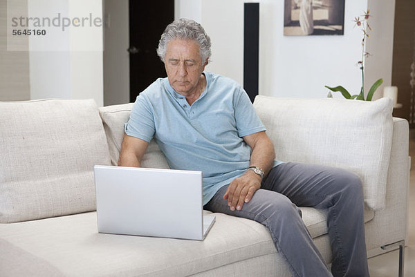 Ein älterer Mann  der auf einer Couch sitzt und einen Laptop benutzt.