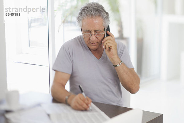 Ein älterer Mann  der das Telefon benutzt und auf ein Dokument schreibt.