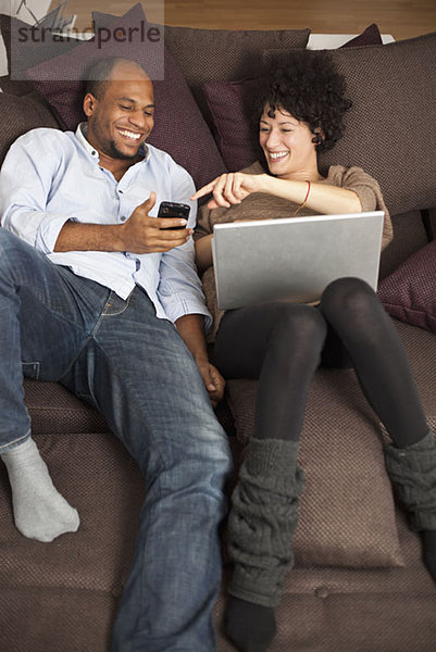 Ein Paar  das auf einem Sofa liegt und über etwas auf einem Handy lacht.