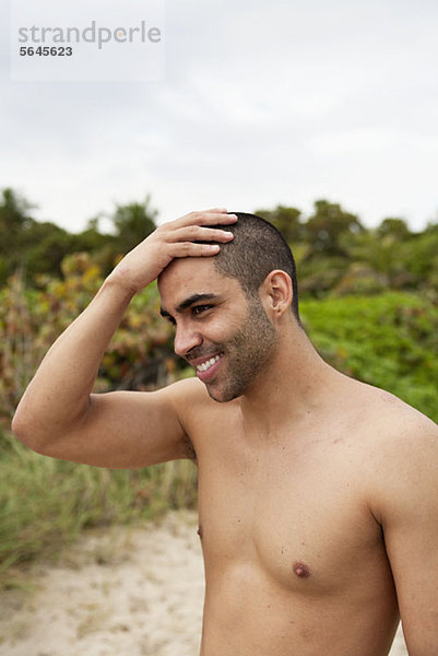 Ein fröhlicher junger Mann reibt sich den Kopf am Strand.