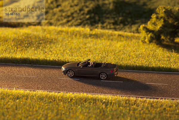 Ein Diorama eines Miniatur-Mannes  der auf einer Landstraße einen Spielzeug-Cabrio-Sportwagen fährt.