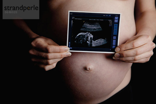 Eine schwangere Frau hält einen Ultraschall vor ihren nackten Bauch.