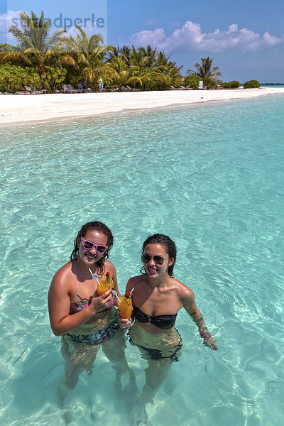 Zwei Mädchen  ca. 14 und 18 Jahre  mit Sonnenbrillen  trinken Cocktails in einer türkisfarbenen Lagune im Meer  hinten Malediveninsel  Malediven  Indischer Ozean  Asien