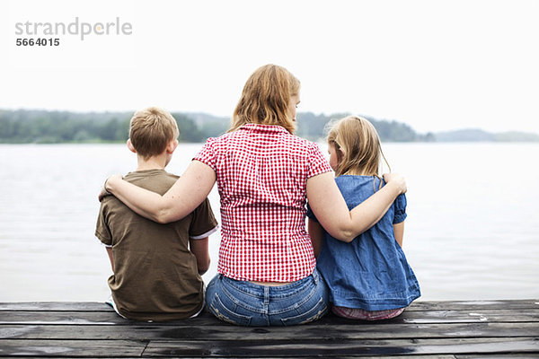 Rückansicht der Mutter mit Arm um ihre Kinder auf dem Pier sitzend