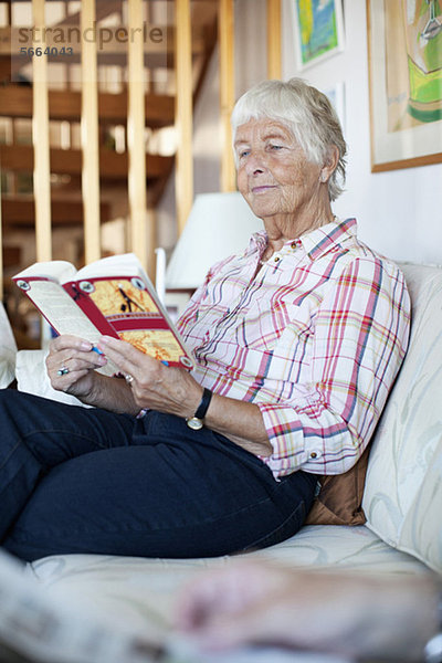 Ältere Frau beim Lesen eines Romans auf dem Sofa im Wohnzimmer